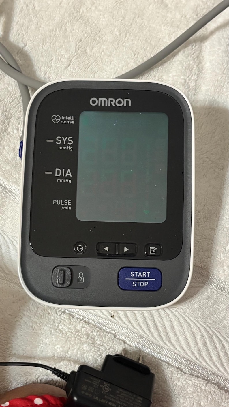 May include: An Omron blood pressure monitor with a black and white casing, a large digital display screen, and a blue Start/Stop button. The device features labels for SYS mmHg, DIA mmHg, and PULSE/min, along with navigation and memory buttons.