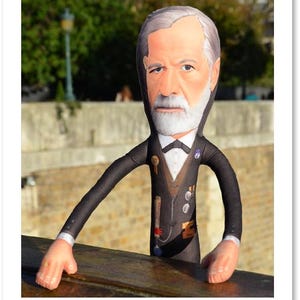 May include: A plush doll of a man with a white beard and gray hair, wearing a black suit with a bow tie and medals. The doll has long arms and is leaning on a wooden surface. The background shows a blurred outdoor scene.
