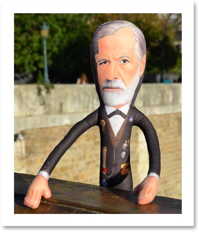 May include: A plush doll of a man with a white beard and gray hair, wearing a black suit with a bow tie and medals. The doll has long arms and is leaning on a wooden surface. The background shows a blurred outdoor scene.