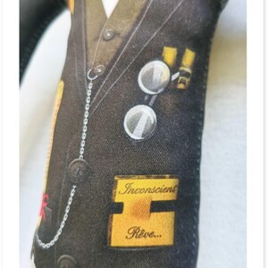 May include: A fabric doll dressed in a black suit with a white shirt and vest. The suit has various patches and a pocket watch chain. The text "Inconscient R&ecirc;ve..." is visible on a yellow patch.