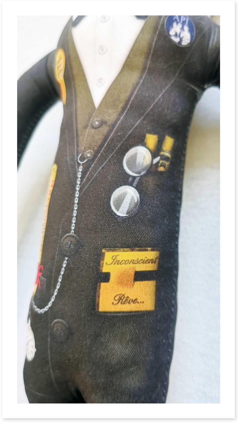 May include: A fabric doll dressed in a black suit with a white shirt and vest. The suit has various patches and a pocket watch chain. The text "Inconscient R&ecirc;ve..." is visible on a yellow patch.