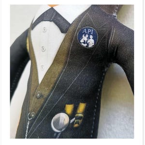 May include: A fabric doll wearing a black pinstripe suit with a white shirt, bow tie, and a pocket watch. The suit has a gold-colored lapel and a small blue pin that says "API".