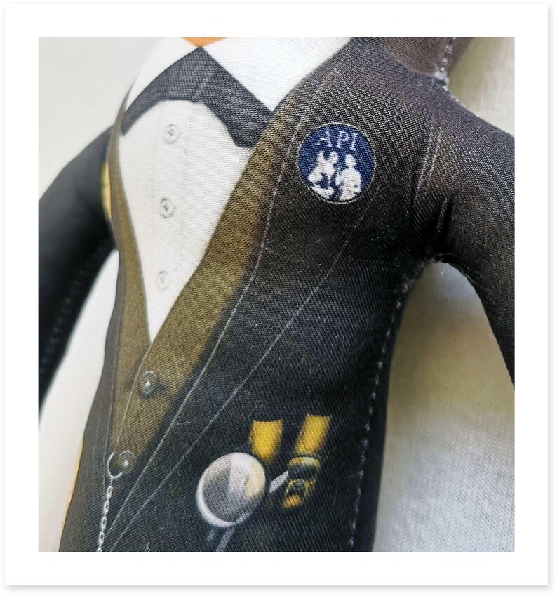 May include: A fabric doll wearing a black pinstripe suit with a white shirt, bow tie, and a pocket watch. The suit has a gold-colored lapel and a small blue pin that says "API".