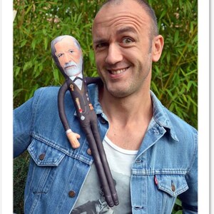 May include: A smiling person wearing a blue denim jacket with a doll of a man with a white beard and a black suit. The doll is draped over the person's shoulder. The background is green foliage.