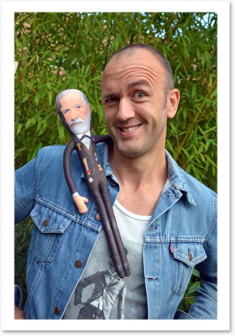May include: A smiling person wearing a blue denim jacket with a doll of a man with a white beard and a black suit. The doll is draped over the person's shoulder. The background is green foliage.