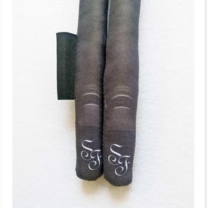 May include: Two black fabric tubes with white cursive "F" initials at the bottom. The tubes have a textured appearance and are positioned vertically. A black rectangular piece of fabric is visible to the left.