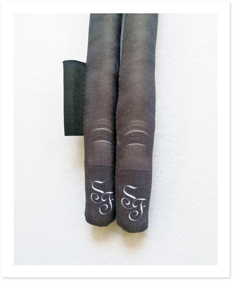 May include: Two black fabric tubes with white cursive "F" initials at the bottom. The tubes have a textured appearance and are positioned vertically. A black rectangular piece of fabric is visible to the left.
