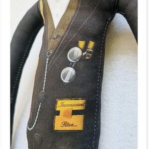 May include: A fabric doll dressed in a black suit with a white shirt and bow tie. The suit has a pocket watch chain, round glasses, and a patch that says "API". A yellow label reads "Inconscient R&ecirc;ve..."