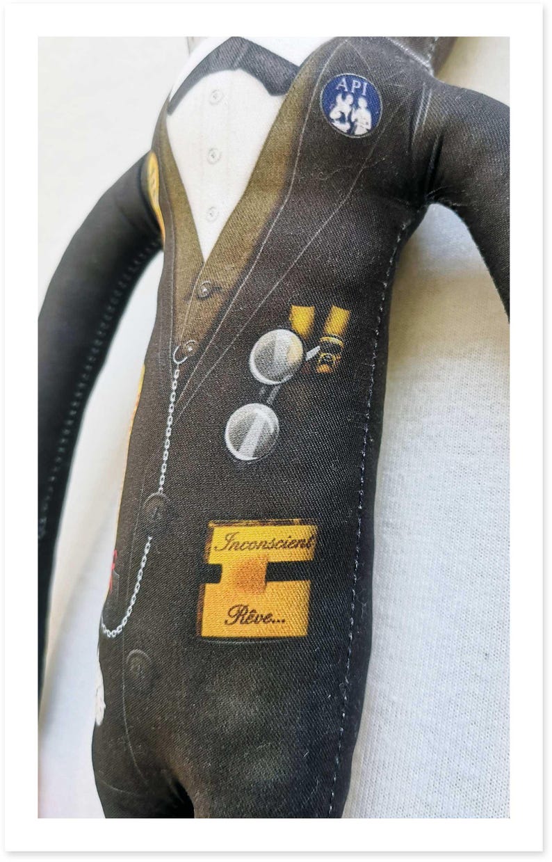 May include: A fabric doll dressed in a black suit with a white shirt and bow tie. The suit has a pocket watch chain, round glasses, and a patch that says "API". A yellow label reads "Inconscient R&ecirc;ve..."