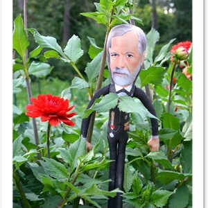 May include: A doll of a man with a gray beard and hair, wearing a black suit and bow tie, stands among green plants and red flowers. The background features a blurred view of trees and foliage, creating a natural setting.