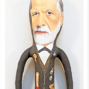 May include: A fabric doll depicting a distinguished man with a white beard and hair, wearing a dark suit with a bow tie. The doll has several pins and a scroll with text. The doll's face is painted with realistic features.