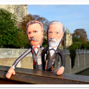 May include: Two plush dolls of historical figures, one with a large mustache and red bow tie, the other with a white beard, stand on a wooden railing. They are dressed in suits with medals, set against a backdrop of a stone building and trees.