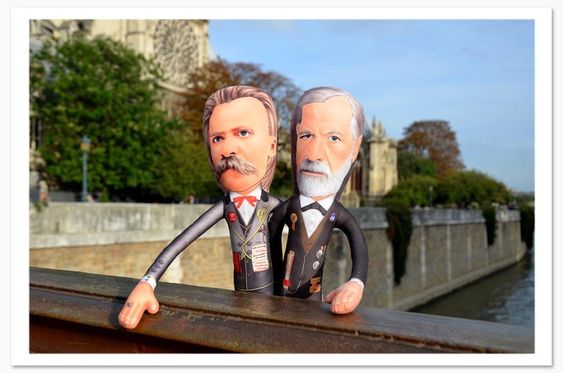 May include: Two plush dolls of historical figures, one with a large mustache and red bow tie, the other with a white beard, stand on a wooden railing. They are dressed in suits with medals, set against a backdrop of a stone building and trees.