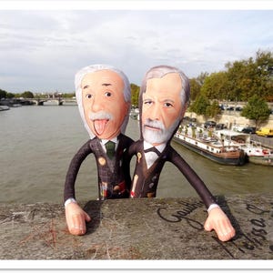 May include: Two plush dolls of famous figures, one with his tongue out, the other with a serious expression, stand on a bridge. They are dressed in suits and ties. The background shows a river, boats, and buildings under a cloudy sky.