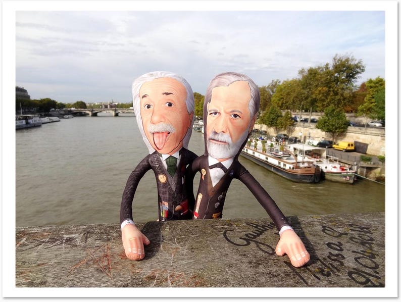 May include: Two plush dolls of famous figures, one with his tongue out, the other with a serious expression, stand on a bridge. They are dressed in suits and ties. The background shows a river, boats, and buildings under a cloudy sky.