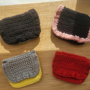 Peut inclure: Quatre portefeuilles crochetés à la main, noirs, noirs avec bordure rose, gris avec base jaune et rouge. Chaque portefeuille a une forme rectangulaire avec un bord à volants et une fermeture à rabat. Présentés sur une surface en bois.