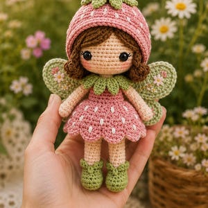 Può includere: Bambola a uncinetto a forma di fata, con cappello e vestito a forma di fragola rosa, ali, colletto e scarpe verdi. La bambola ha i capelli castani e gli occhi neri. Lo sfondo è sfocato con fiori.