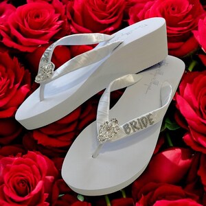 May include: White wedge flip-flops with satin straps and rhinestone embellishments. One flip-flop features the word "BRIDE" in silver rhinestones. The background is a bed of red roses, creating a romantic theme.