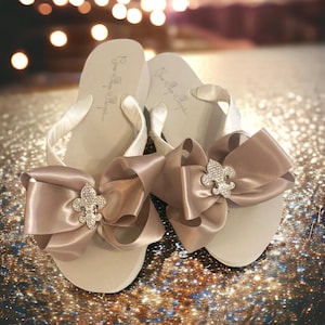 May include: A pair of beige flip-flops with white straps and large, satin bows in a matching color. Each bow features a silver fleur-de-lis embellishment. The flip-flops are on a glittery surface.