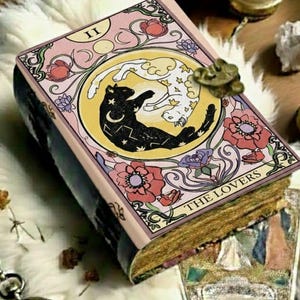 Puede incluir: Un diario temático de cartas del tarot con una cubierta rosa que presenta un diseño de gato blanco y negro dentro de un círculo amarillo. El diario está etiquetado como "THE LOVERS" y tiene un cierre decorativo. También se ven una vela y un reloj de bolsillo.