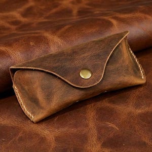 May include: A brown leather glasses case with a snap closure. The case is made of distressed leather and has a stitched edge. The case is resting on a textured brown leather surface.