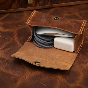 May include: Open brown leather pouch with a snap closure, holding a white mouse, a gray cable, and a white charging block. The pouch is made of distressed leather, and the background is a similar leather surface.