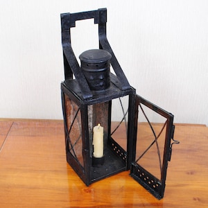 Op de afbeelding: Een zwarte metalen lantaarn met een rechthoekig frame, een scharnierende deur en een handvat aan de bovenzijde. De lantaarn heeft een cilindervormige schoorsteen en heldere glazen ruiten. Binnenin brandt een crèmekleurige stompkaars.