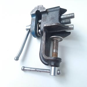 May include: A metal bench vise featuring a dark gray cast iron body with a blue accent on the side. The tool includes a threaded screw mechanism with a silver handle for tightening, a clamp base for attachment, and two parallel metal guide rods.