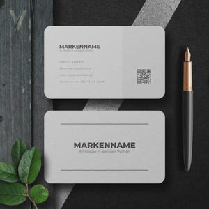 Peut inclure: Deux cartes de visite rectangulaires gris clair aux coins arrondis. La carte du haut affiche MARKENNAME, des coordonnées et un QR code. La carte du bas présente MARKENNAME et un slogan. Un stylo plume noir et doré est placé à droite.
