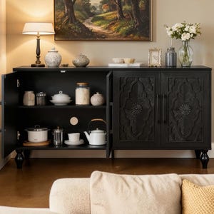 May include: A black wooden sideboard with carved floral designs on the doors. The cabinet is open, revealing shelves with various kitchen items, including jars, dishes, and a white teapot. A lamp and artwork are on top.