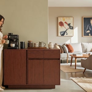 May include: A dark brown wooden sideboard featuring a single door on the left and two drawers above two doors on the right. The sideboard surface holds a black espresso machine, a black pitcher, a glass jar, ceramic mugs, and a ribbed vase.