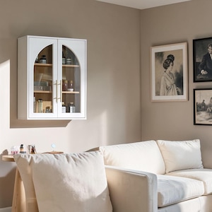 May include: A white wall-mounted cabinet with two glass doors featuring arched tops and vertical gold-toned metal handles. The interior contains wooden shelves holding various small bottles and containers, visible through the clear glass panels.