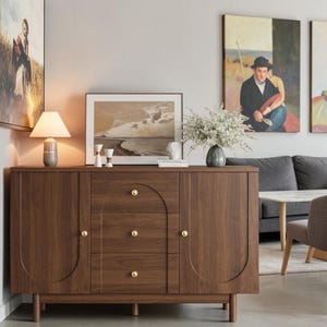 May include: A dark brown wooden sideboard featuring two side cabinets and three central drawers. Each drawer and cabinet door is accented with a small, round, gold-colored knob. The front panels display a subtle arched design detail.