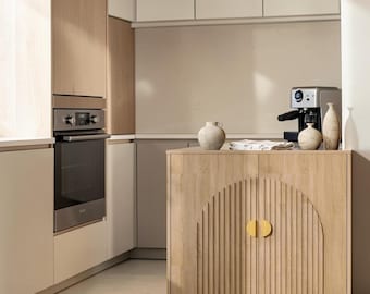 May include: A light wood kitchen island cabinet featuring a central arched design with vertical slats and two semi-circular gold handles. The cabinet is topped with three textured beige ceramic vases and a folded white cloth.