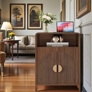 May include: A dark wood cabinet with a curved top, featuring vertical fluted detailing on the two doors and two semi-circular gold handles. The open top shelf holds a tablet, a pair of headphones, and two books titled Kinfolk and LIKEtoKNOWit.