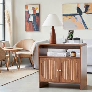 May include: A wooden nightstand with a smooth top surface, an open shelf, and two cabinet doors featuring vertical slats within circular frames. The cabinet has small round gold knobs and sits on a flat base. A lamp and phone charger sit on top.