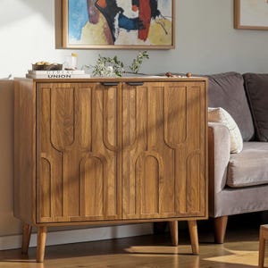 May include: A wooden cabinet with two doors and a mid-century modern design. The cabinet is made of light brown wood and features a decorative arched pattern on the doors. It has four tapered legs and black handles. The cabinet is placed next to a gray sofa.