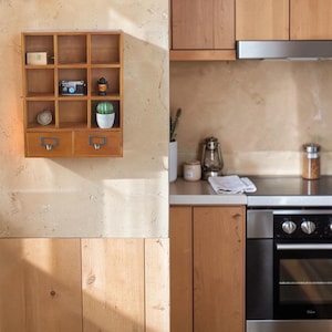 May include: A wall-mounted wooden display shelf featuring nine open cubby compartments and two bottom drawers with metal label pulls. The shelves contain a small wooden box, a miniature camera, a lantern, a ball of twine, and a potted cactus.