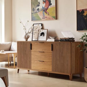May include: A light brown wooden sideboard featuring a central column of three horizontal drawers with thin black handles, flanked by two side cabinets with vertical slatted doors and black vertical handles, all supported by four tapered legs.