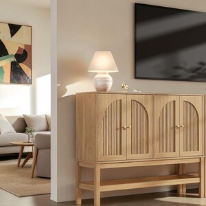 May include: A light oak wood sideboard featuring four cabinet doors with vertical slatted detailing and arched tops. The cabinet has small round gold knobs, a lower open shelf, and stands on four legs. A white table lamp sits on the top surface.