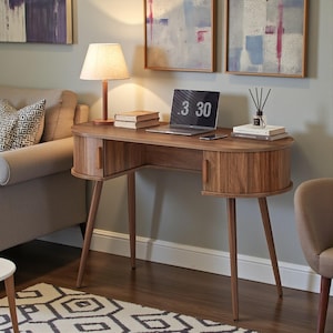 May include: A mid-century modern wooden desk with a curved design, two small cabinets, and tapered legs. A laptop displays "3 30" on the screen. A lamp, books, and a phone are on the desk. The desk is in a room with artwork and a sofa.