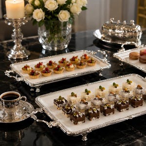 Puede incluir: Una mesa con bandejas plateadas que contienen varios postres, incluyendo tartaletas y macarons. También hay una taza de café y un jarrón con rosas blancas. La mesa tiene una superficie de mármol negro.