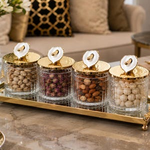 Op de afbeelding: Een rechthoekig spiegelplateau met een gouden metalen frame en decoratieve bloemaccenten op de hoeken, met vier heldere glazen potten. Elke pot heeft een glazen behuizing met textuur, een gehamerd gouden deksel en een handvat in de vorm van een calla-lelie.