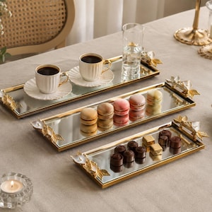 May include: Three rectangular mirrored trays with gold-toned metal frames and decorative floral handles at each end. The trays are arranged in a tiered formation, displaying two white coffee cups on saucers, colorful macarons, and assorted chocolates.