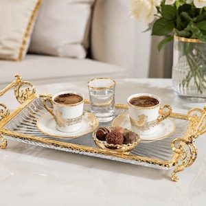 以下が含まれることがあります： 2つの白と金のコーヒーカップ、ソーサー、チョコレートの小さなボウルが付いた装飾的なサービングトレイ。水と白い花の入った花瓶も付いています。トレイは金色の透かし彫りのデザインです。