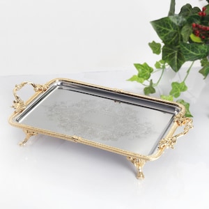 May include: A rectangular silver serving tray with ornate gold-coloured trim, handles, and feet. The tray has a decorative floral pattern in the centre. The tray is on a white surface.