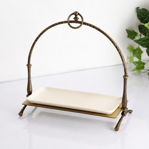 May include: A rectangular white ceramic tray set within a bronze-toned metal stand. The stand features a high, arched handle with a circular ring at the top and four small, curved feet that support the base plate holding the ceramic tray.