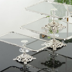 May include: A three-tier serving stand featuring rectangular glass plates with silver-toned, rope-textured edges. The stand is supported by ornate, metallic silver bases with intricate detailing and clear, faceted crystal-like accents at each level.