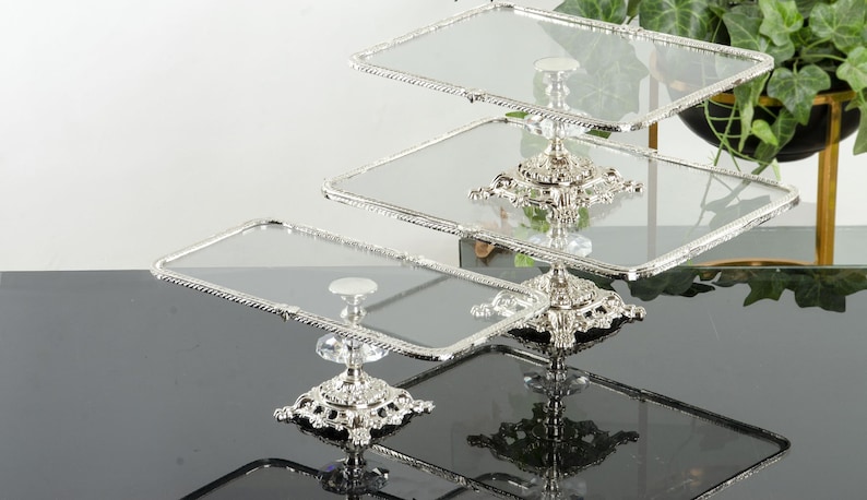 May include: A three-tier serving stand featuring rectangular glass plates with silver-toned, rope-textured edges. The stand is supported by ornate, metallic silver bases with intricate detailing and clear, faceted crystal-like accents at each level.