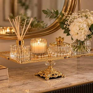 May include: A rectangular gold-rimmed mirrored tray with a decorative pedestal base. The tray holds a glass reed diffuser with wooden sticks, a lit candle in a ribbed glass holder, a lidded glass jar with gold accents, and a vase of white flowers.
